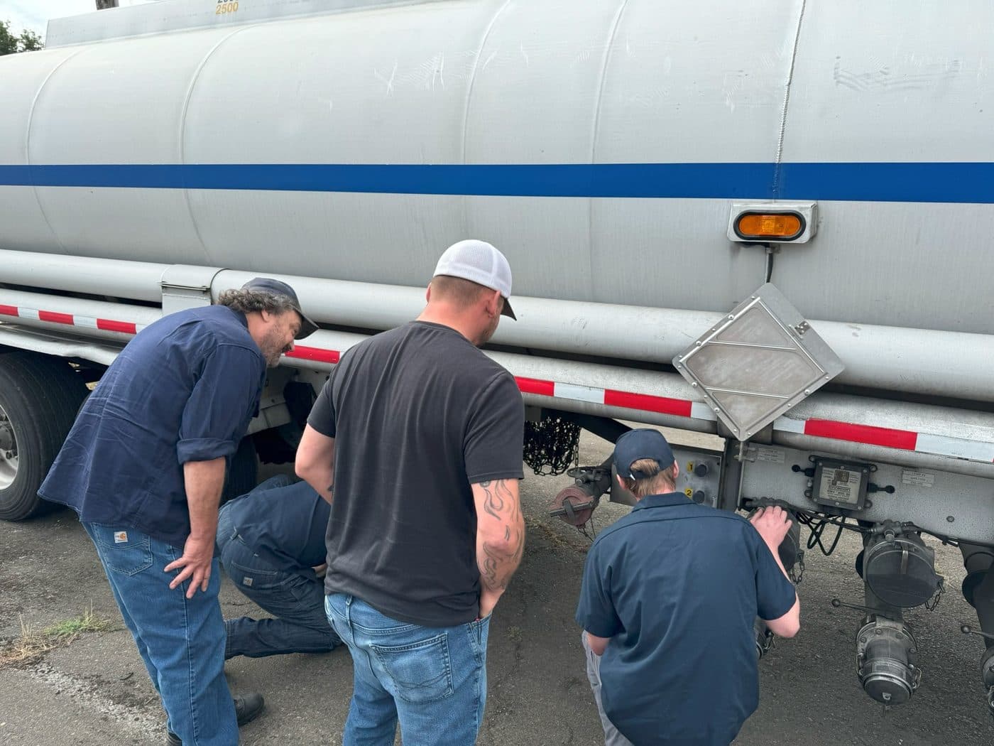 Three inspectors crouched at the rear of a specification cargo tank trailer, examining the piping and the placard holder